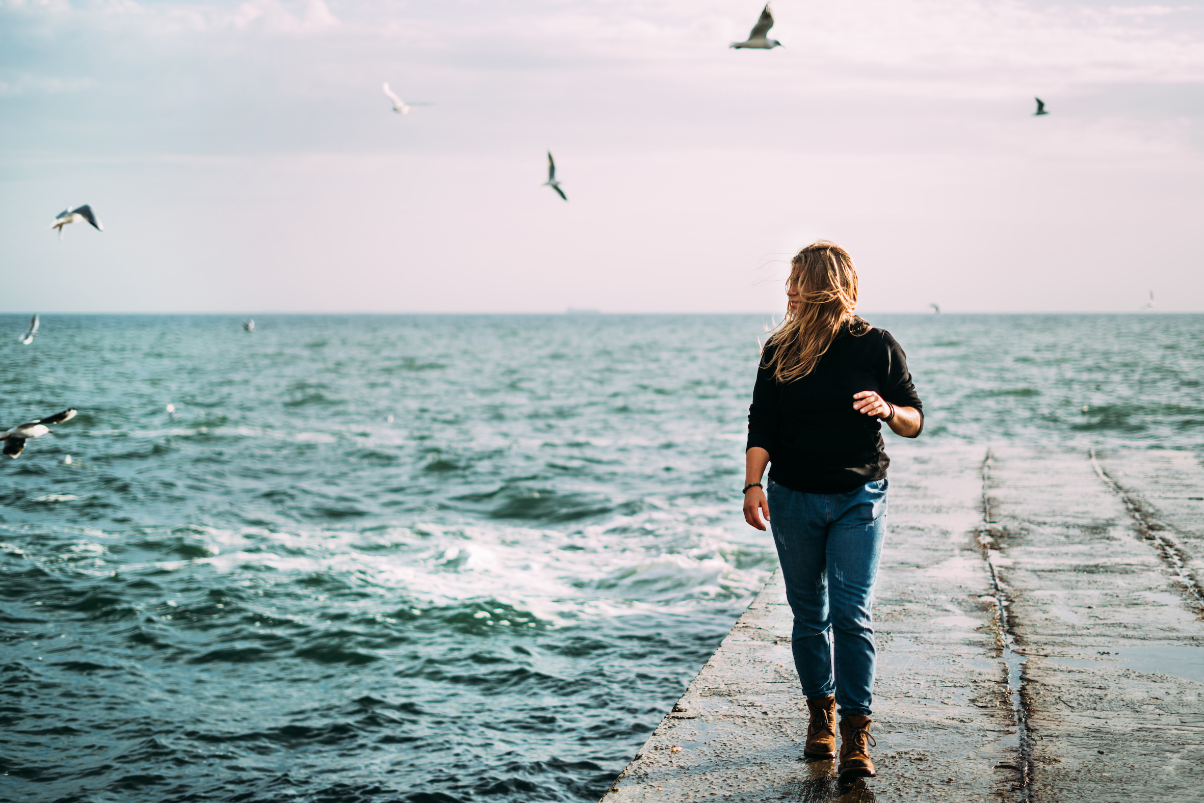 En tjej går på en brygga vid havet med vågor och hård vind i svart tröja och jeans. Rosa gryning och måsar på himlen.