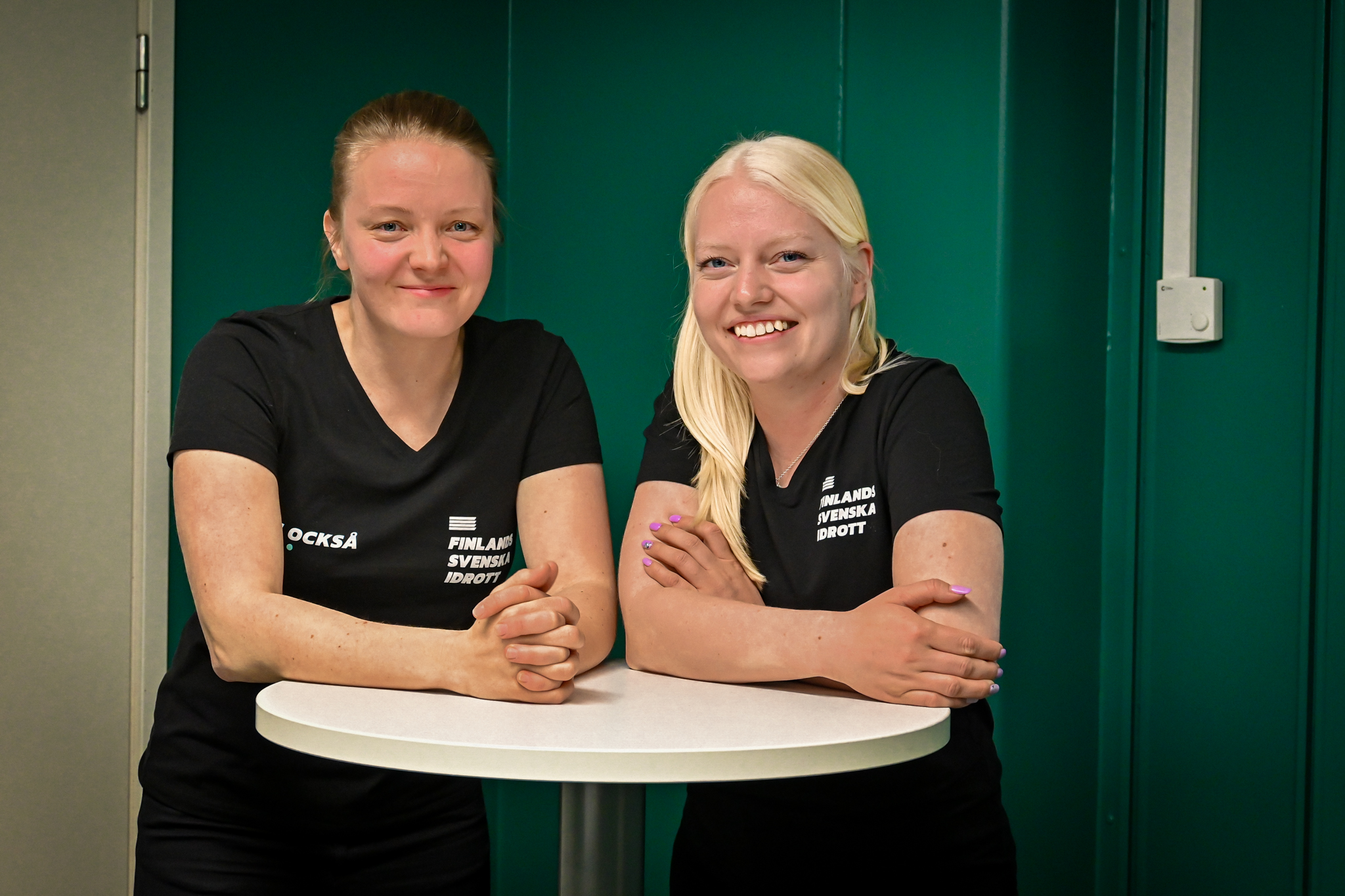 Lotta Nylund och Tarja Krum står bredvid varandra vid ett vitt bord mot en grön vägg.