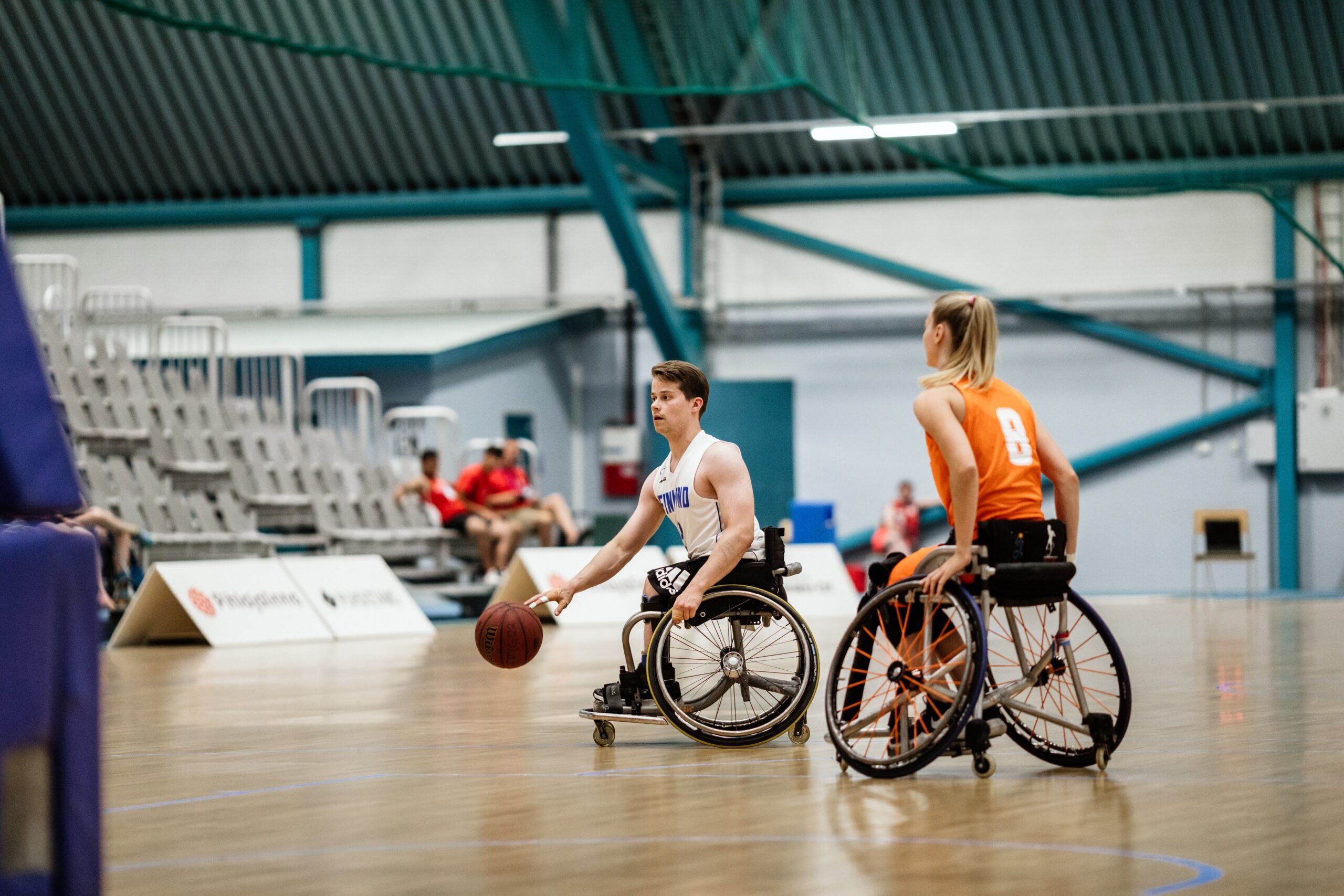 Två spelare i rullstol som spelar korgboll inomhus.