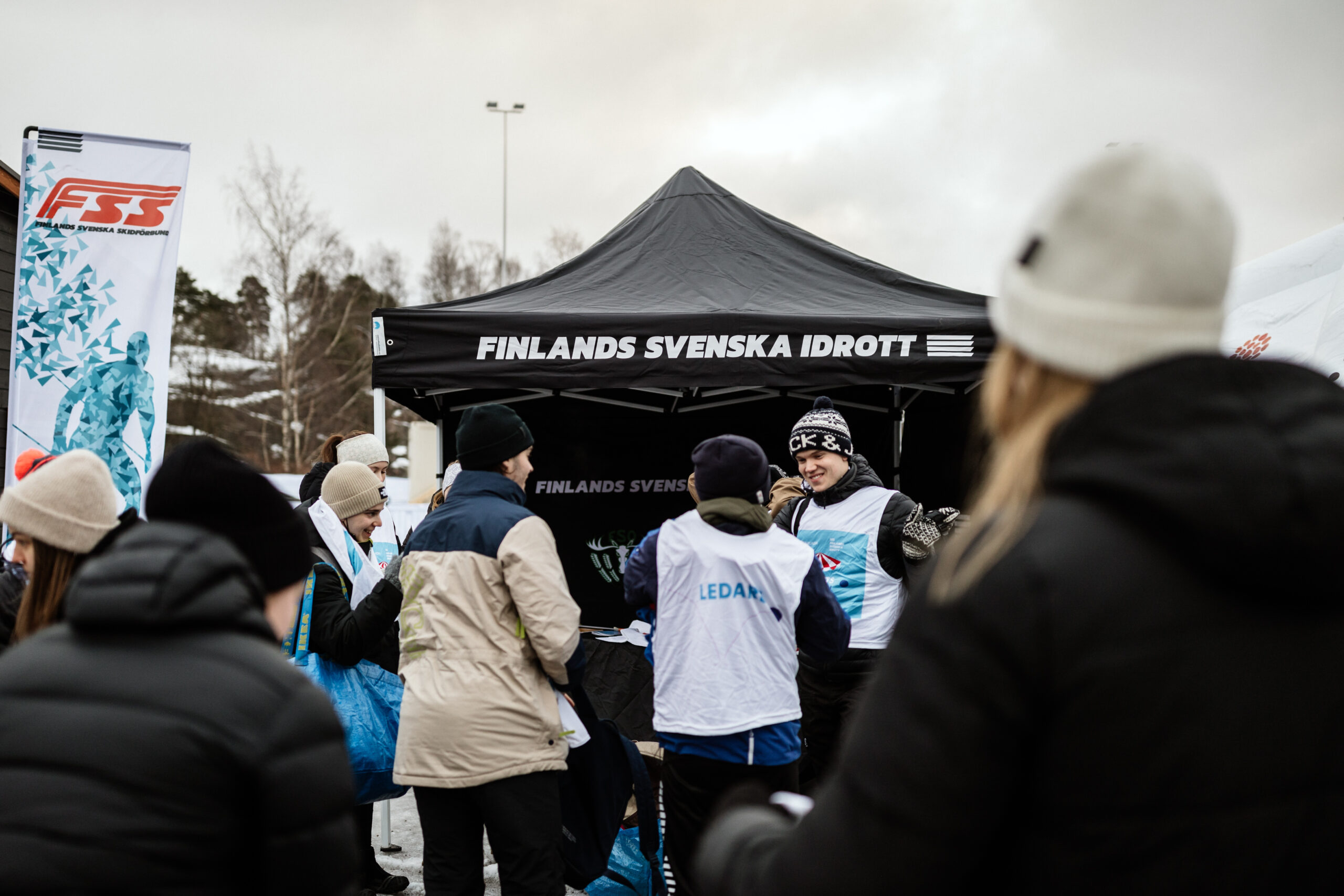 Människor står samlade ute och hälsar glatt på varandra. I bakgrunden kan man se en FSS flagga och ett tält med texten Finlands Svenska Idrott.