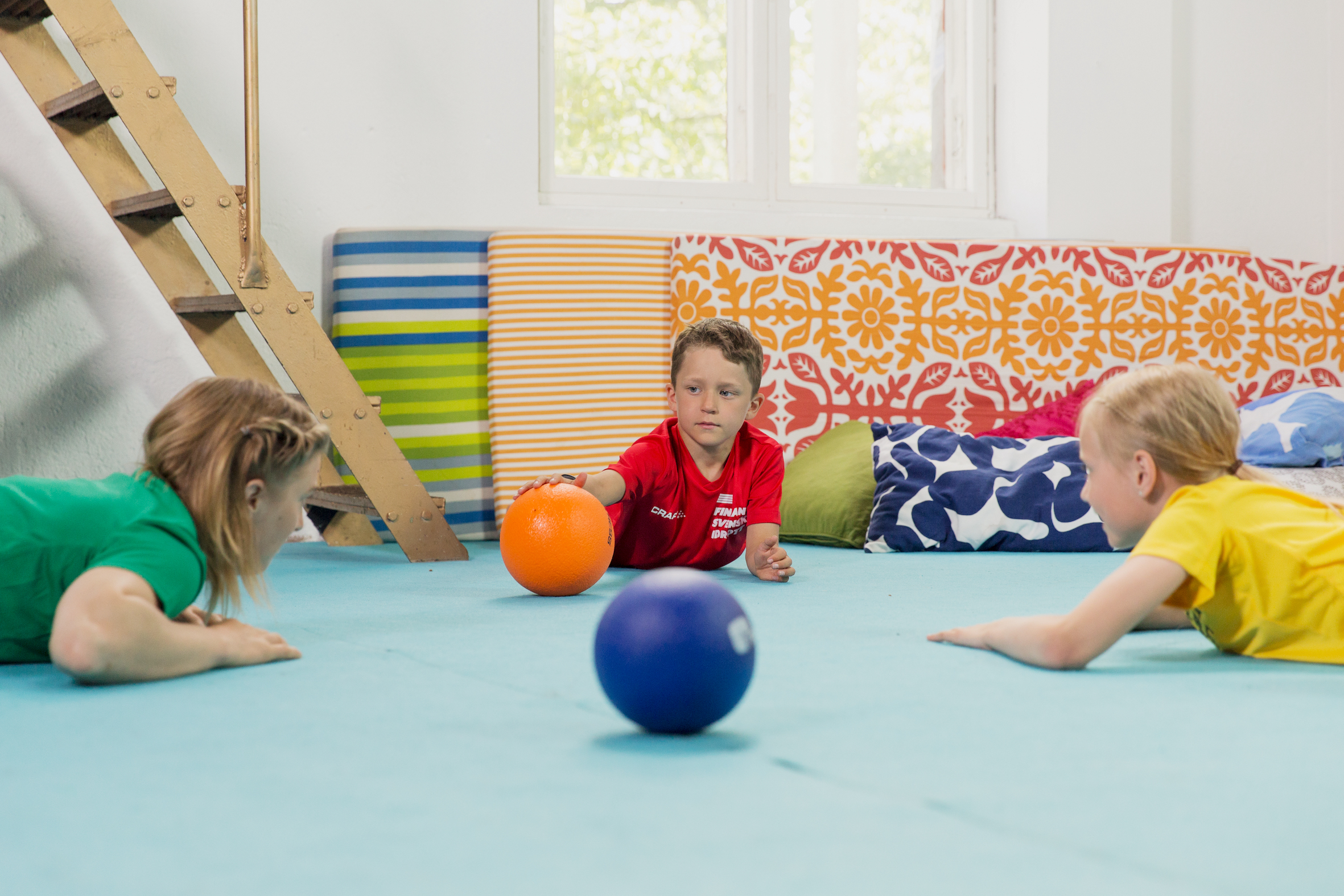 En vuxen och två barn ligger runt varandra och rullar bollar mot varandra inne i en gymnastiksal.