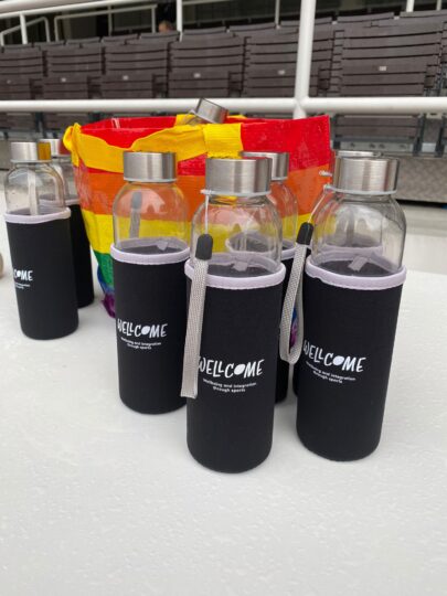 Sju vattenflaskor i glas där det står WELLcome. I bakgrunden finns en regnbågsfärgad påse med mera flaskor.